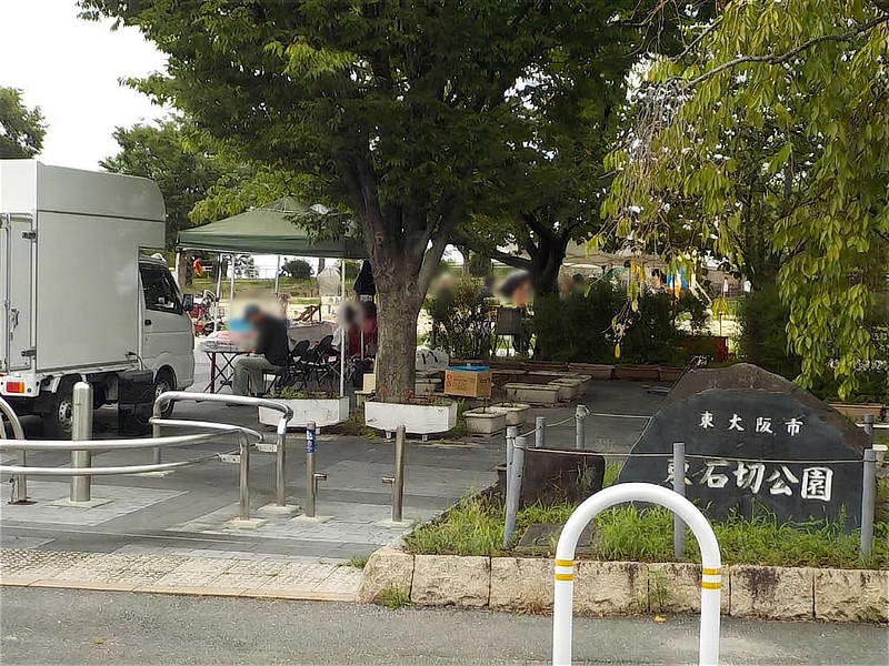 東大阪市 東石切公園の石碑がある入口付近。左側にトラックと緑のテントがあり、木陰で出店者が準備や販売をしている様子。