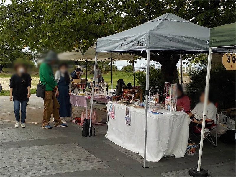 灰色のテントの下でハンドメイドアクセサリーや雑貨を販売する店舗。来場者が商品を選び、出店者とやり取りをしている様子。