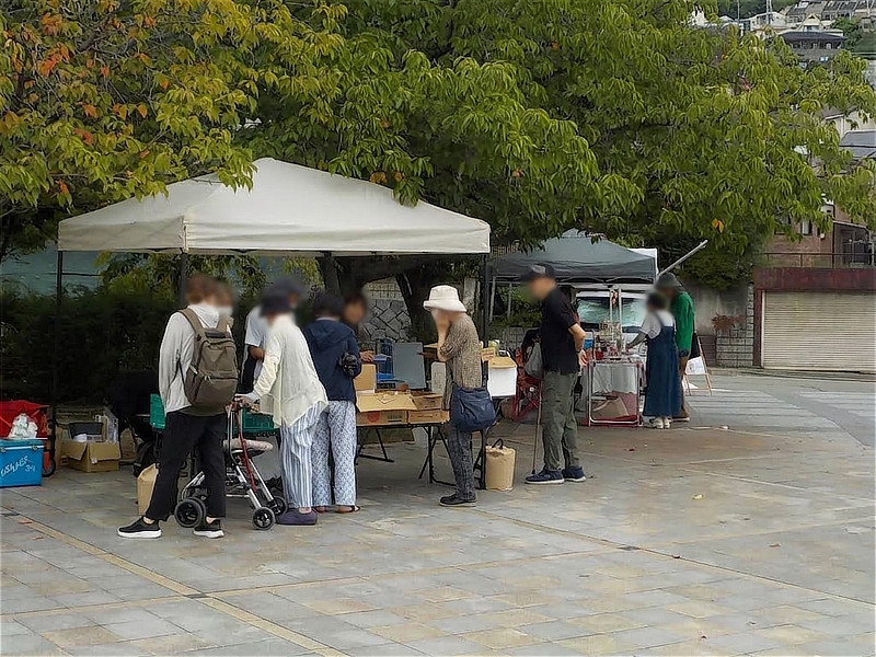 大きな木の下に設営された店舗で買い物をする来場者たち。歩行補助車を押した高齢の方々が商品を眺めている。